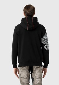 Schwarzer Hoodie mit roten Nähakzenten, versehen mit einem großen Grafikdesign am linken Ärmel; kombiniert mit lässigen, hellen Denim-Jeans.