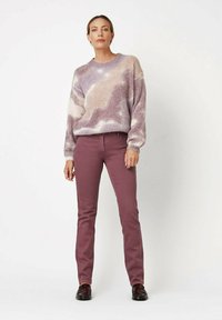 Hellvioletter, strukturierter Pullover mit einem marmorierten Muster, kombiniert mit figurbetonten, burgunderfarbenen Chinos. Das Modell trägt dunkle Schuhe und steht vor einem schlichten Hintergrund.