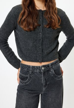 Femme portant un cardigan court gris foncé boutonné et un jean noir délavé taille haute, aux longs cheveux bruns ondulés.