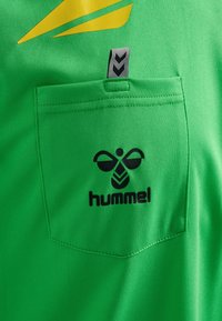 Maillot de sport vert avec une finition texturée, présentant un logo "hummel" noir, une poche et un élément de design diagonal jaune.