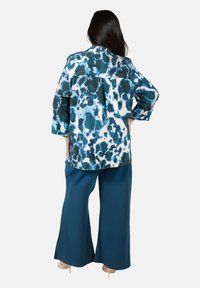 Luftige, mehrfarbige Bluse mit einem blau-weißen abstrakten Muster, kombiniert mit türkisfarbenen, weiten Hosen. Glatte Stofftextur.