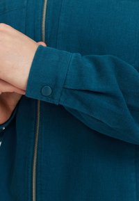 Teal corduroy jas met een ritssluiting, met een gestructureerde oppervlakte en een drukknoopdetail op de manchet. Close-up van de mouw.