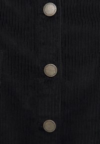 Zwart corduroy stof met verticale ribbels, voorzien van vier zilveren knopen met merknaam, geplaatst langs de voorflap.