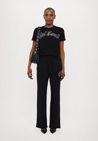 Juodas marškinėlis su "Just Cavalli" sidabriniu raštu, derinamas su juodomis kelnėmis su raišteliais. Papildomi aksesuarai - maža juoda rankinė ir smailiakulnės bateliai.