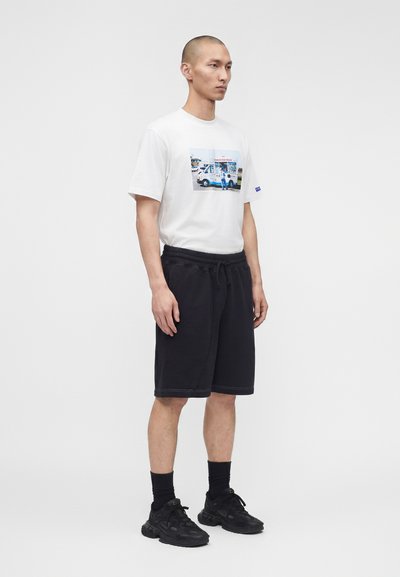 Άνδρας φοράει λευκό T-shirt με σχέδιο, μαύρα σορτς με κορδόνι, μαύρες κάλτσες και μαύρα αθλητικά παπούτσια, στέκεται μπροστά σε ανοιχτόχρωμο απλό φόντο.