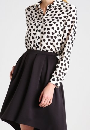 Kobieta ubrana w białą, zapinaną na guziki bluzkę z nieregularnymi czarnymi plamami oraz czarną plisowaną spódnicę z wysokim stanem na jednolitym tle.