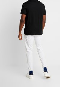 Černé tričko s krátkým rukávem v kombinaci s bílými slim-fit kalhotami. Bílé tenisky s námořnickými akcenty; model otočený zády k fotoaparátu.