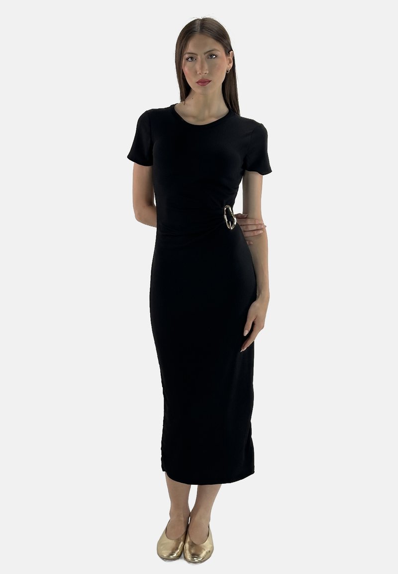 Das Model posiert selbstbewusst in einem figurbetonten schwarzen Kleid mit einem Gürteldetail, trägt goldene Ballerinas und minimalen Schmuck vor einem schlichten Hintergrund.