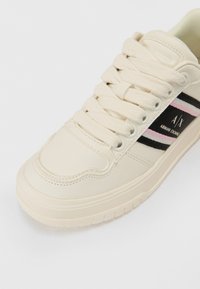 Kremfargede sneakers med en glatt lærfinish, med horisontale svarte og rosa striper og en logo på siden. Snøring.