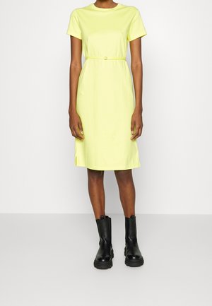 Personne portant une robe jaune à manches courtes, longueur genou, avec une ceinture fine, associée à des bottines noires épaisses, debout sur un fond uni.