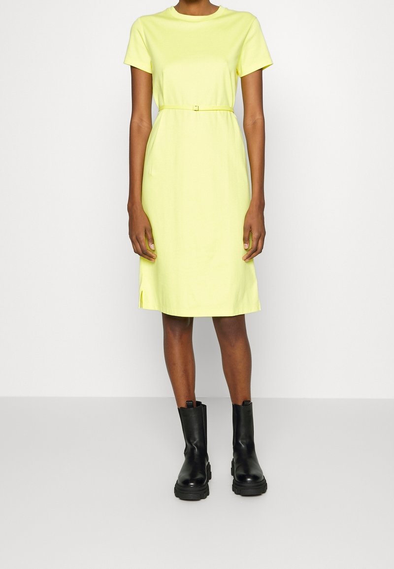 Personne portant une robe jaune à manches courtes, longueur genou, avec une ceinture fine, associée à des bottines noires épaisses, debout sur un fond uni.