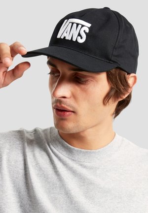 Junger Mann, der eine schwarze Vans-Kappe justiert, trägt ein hellgraues Sweatshirt und schaut nach unten vor einem einfarbigen grauen Hintergrund.