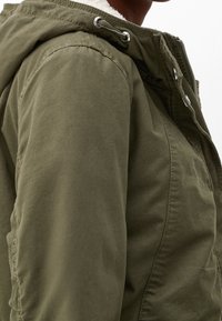 Olivově zelená bunda s texturovanou, odolnou tkaninou. Má zip, patentky, kapuci se stahovacími šňůrkami a měkkou vnitřní podšívku.
