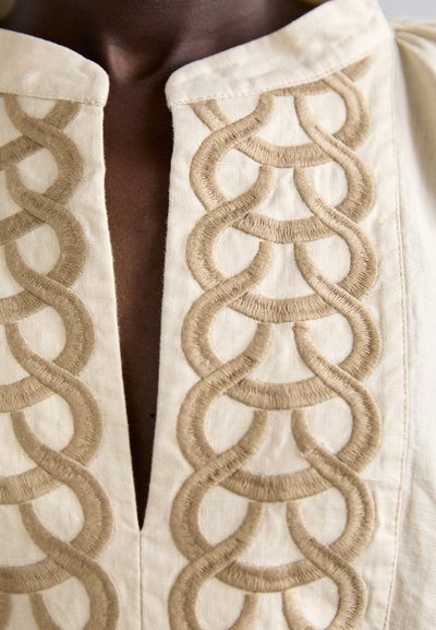 Un torse à la peau foncée arborant une chemise beige ornée d'un motif brodé en boucle beige le long du col et de l'ouverture sur le devant.