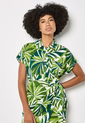 Donna con capelli naturalmente ricci che indossa un abito verde e bianco con motivo a foglie, maniche corte e bottoni, in piedi con una mano sul fianco.