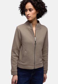 Femme aux cheveux courts et bouclés portant une veste beige texturée à fermeture éclair et un jean bleu foncé, regardant vers sa gauche sur un fond uni.