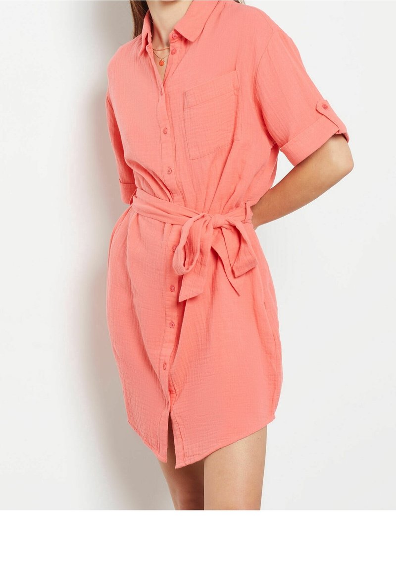 Femme portant une robe-chemise corail à manches retroussées et une ceinture nouée à la taille, debout devant un fond blanc.