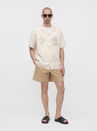 Homme portant une chemise crème à manches courtes avec des motifs, un short beige, des sandales noires et des lunettes de soleil, debout devant un fond blanc uni.