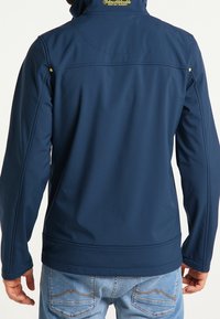 Marinblå softshell-jacka med hög krage, sömma detaljer och gult logotyp på baksidan. Har stretchigt material och figursydd design.