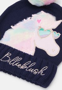 A tengerészkék kötött sapka pasztell unikornis mintával rendelkezik, amelynek texturált, színes sörénye, szív alakú szemei vannak, és ezüst színű "Billieblush" hímzés díszíti.
