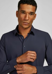 Donkerblauw, lang mouw shirt van gladde stof, met een knoopsluiting aan de voorkant en puntige kraag. De manchet heeft knoopaccenten.