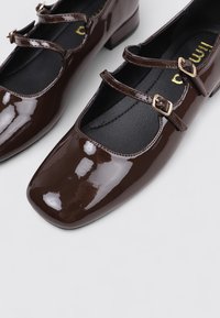 Zapatos Mary Jane de charol marrón con dos correas ajustables y hebillas doradas, punteras cuadradas y tacones bajos.