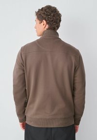 Ruda pūkuota striukė su aukštu apykakle, pasižyminti tekstūruotu audiniu, ilgomis rankovėmis ir šiek tiek siaurėjančiu siluetu apačioje.