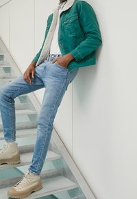 Teal denimjacka med fårahalskrage, vit t-shirt, ljusblå åtsittande jeans och beige snörboots på en trappa.