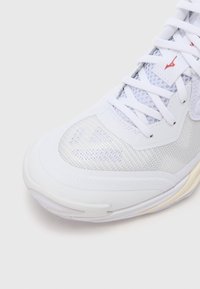 Witte sportschoen met een bovenwerk van mesh en synthetisch materiaal, een afgeronde neus, gestructureerde details en contrasterende rode logo-accenten. Lichtzoolontwerp.