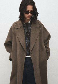 Bruine oversized wollen jas met een brede revers, gedragen over een donker gevoerd gebreid sweater met ruitpatroon en blauwe jeans. Accessoires zijn onder andere donkere zonnebrillen.