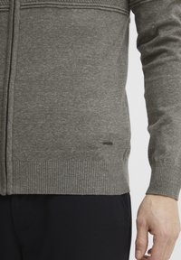 Grå strikket cardigan med ribbestrikkede mansjetter og hem, med et strukturert design og en liten merkevarelogo på siden.