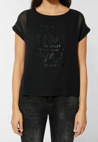 Svart kortärmad topp med rund halsringning, prydd med text av texturiserad metallisk finish på framsidan. Tillverkad av lätt tyg med en avslappnad passform.