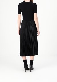 Žena oblečená v černém topu s krátkými rukávy, plisované midi sukni, černých ponožkách a černých botách na podpatku, stojící zády ke kameře.
