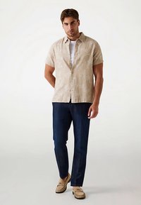 Chemise beige à manches courtes avec un motif discret de feuilles, associée à un pantalon bleu foncé et des chaussures marron clair. Coupe décontractée et confortable.