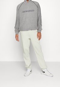 Grå zip-up sweatshirt med texten "LETS FRANS", kombinerad med ljusa sweatpants och vita högskaftade sneakers. Mjuk tygkvalitet och avslappnad passform.