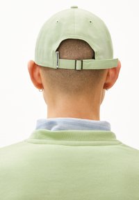 Casquette en coton verte avec une sangle réglable, comportant une petite étiquette à l'arrière. Le sujet porte une chemise bleu clair en dessous.