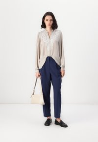 Beige bluse med hvite blonderdetaljer, løs passform, sammen med marineblå bukser og svarte loafers. Holder en liten beige håndveske.