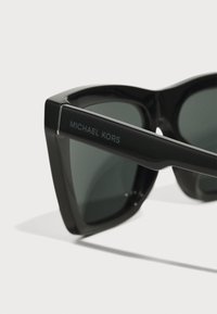 Ochelari de soare negri cu o ramă groasă și unghiulară. Lentile de un verde închis și un logo argintiu pe sprijin. Textură de plastic neted.