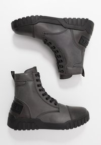 Par de botas de combate de cuero gris oscuro con suelas de goma negra gruesas y cordones, mostradas desde vistas lateral y de la suela sobre un fondo blanco.