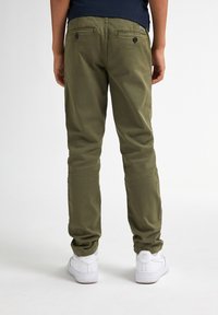 Oliegroene chinos met twee achterzakken, knoopsluitingen en een slim fit, gecombineerd met witte sneakers. De stof lijkt glad en duurzaam.