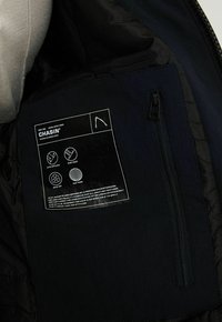 Intérieur de la veste noire montrant une poche zippée et une étiquette détaillant les caractéristiques déperlantes, coupe-vent, séchage rapide et maintien de la chaleur.