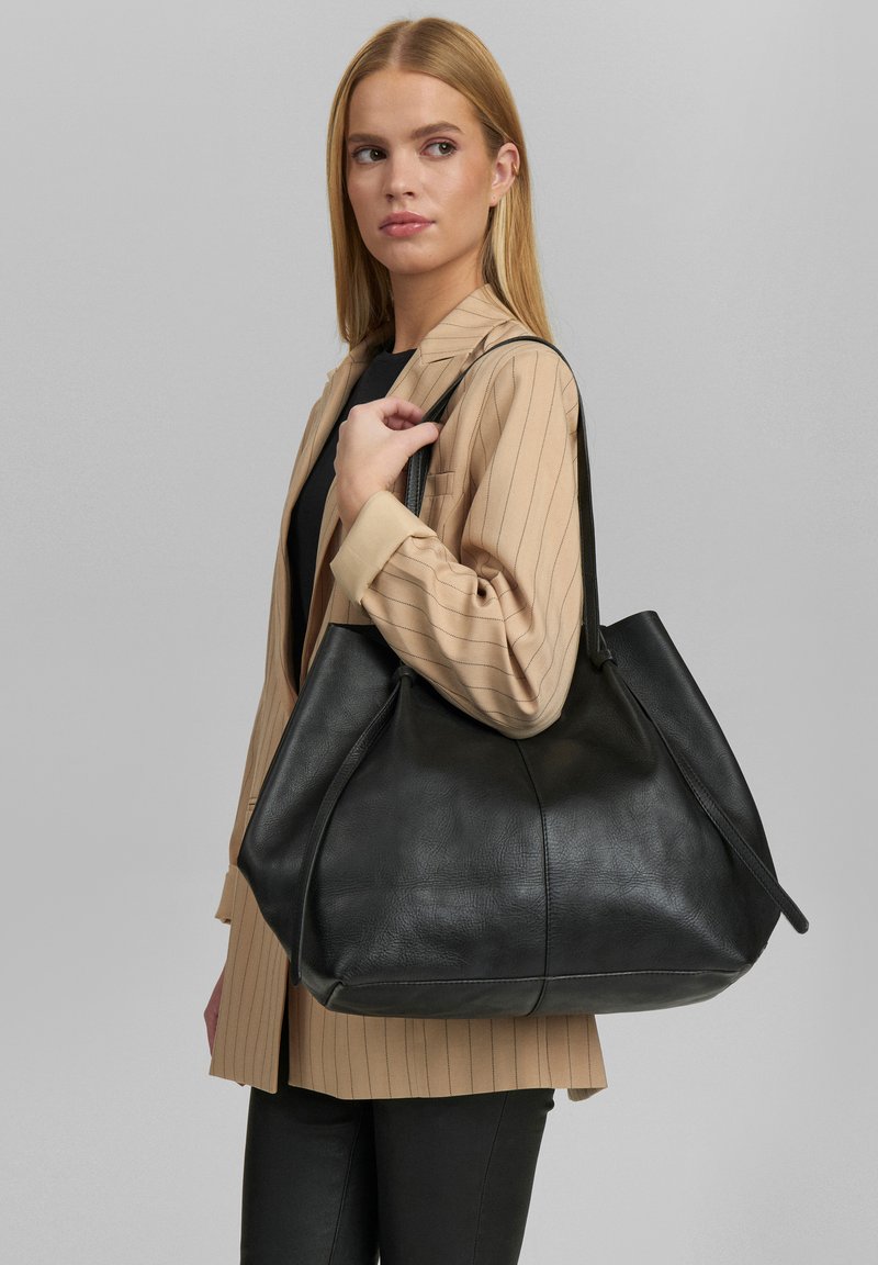 Borsa tote in pelle nera con una finitura morbida, forma arrotondata e due lunghe cinghie per la spalla. Indossata da una persona con un blazer a righe beige.
