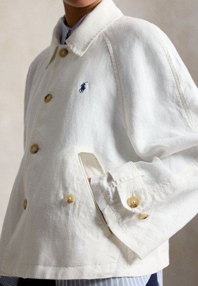 Jachetă albă cu nasturi și logo albastru cu jucător de polo brodat, purtată peste o cămașă cu dungi albastre, mână băgată în buzunarul lateral.