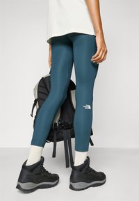 Turkusowe legginsy z białym logo, zestawione z czarnymi butami turystycznymi i skarpetkami sięgającymi do kostek, na tle czarno-szarego plecaka.