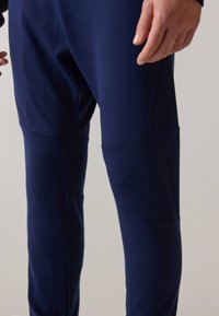 Mørkeblå treningbukser laget av et glatt, elastisk stoff. Egenskaper inkluderer stitched paneler, avsmalnende ben og en slank passform.