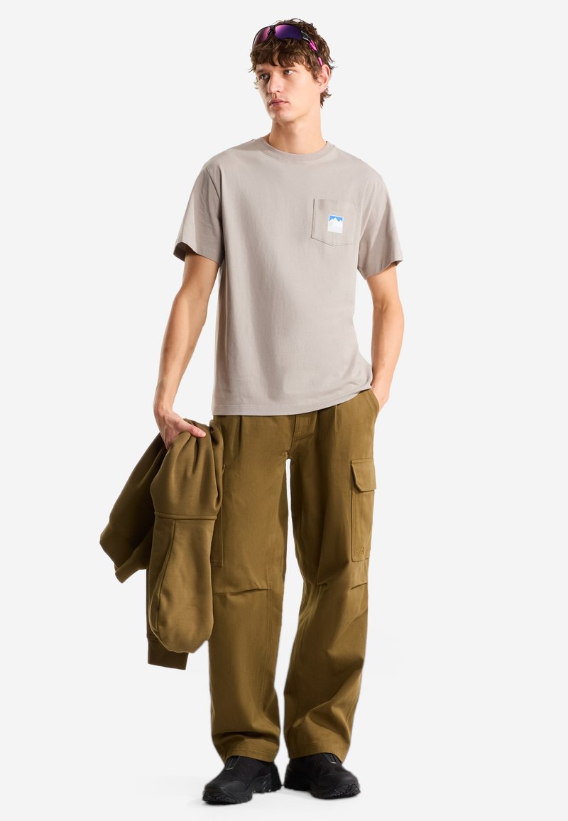 Joven con camiseta color topo y pantalones cargo color oliva, sosteniendo una chaqueta a juego de color oliva, con gafas de sol apoyadas en su cabello rizado.