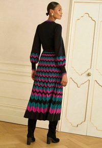 Rochie neagră cu mâneci lungi și bufante; model zigzag colorat în nuanțe de teal, roz și negru. Fusta pliată se extinde până la glezne. Puritată cu cizme negre.