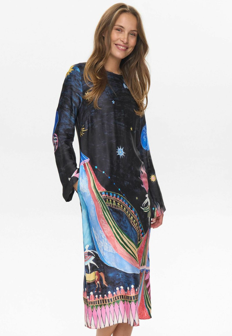 Una mujer sonriente con pelo largo lleva un vestido negro de manga larga con estampados coloridos de temática circense y celeste, de pie frente a un fondo blanco.