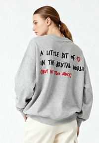 Maglione grigio oversize con scollo rotondo. Sul retro, il testo in nero e rosso recita: "Un po' di ❤️ in questo mondo brutale (ma non troppo)."