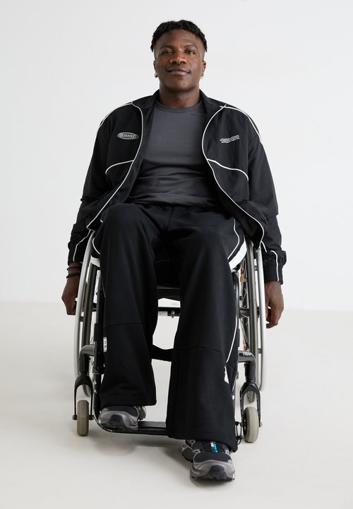 Sportkleding: zwarte jas met witte piping, grijs T-shirt, zwarte losse broek, grijze sneakers. Persoon zittend in een rolstoel.